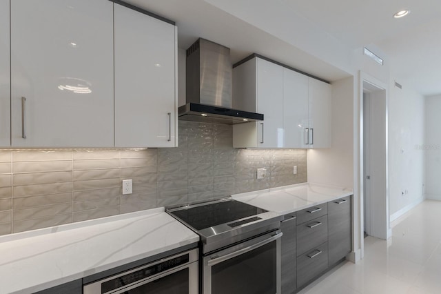 kitchen with tasteful backsplash, wall chimney exhaust hood, white cabinetry, light stone countertops, and stainless steel electric range oven