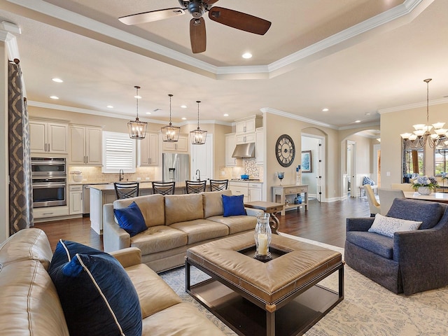 living room with ceiling fan with notable chandelier, light hardwood / wood-style flooring, a raised ceiling, sink, and ornamental molding