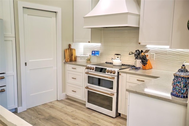 kitchen with light wood-type flooring, tasteful backsplash, white cabinets, double oven range, and premium range hood