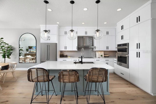 kitchen featuring tasteful backsplash, stainless steel appliances, wall chimney range hood, pendant lighting, and a kitchen island with sink