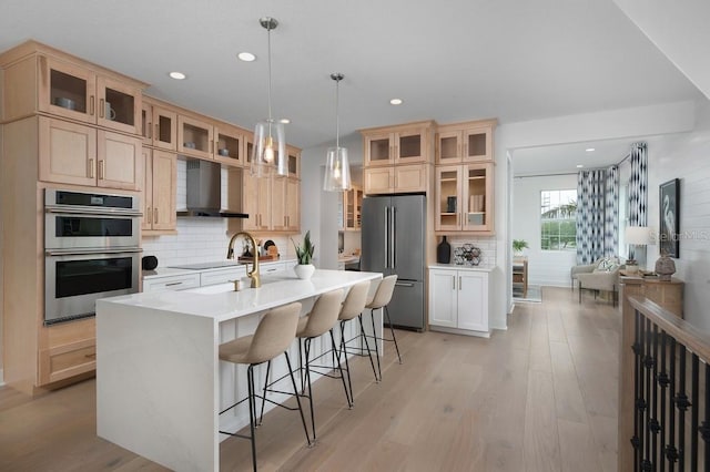 kitchen featuring appliances with stainless steel finishes, tasteful backsplash, a kitchen island with sink, wall chimney range hood, and light wood-type flooring