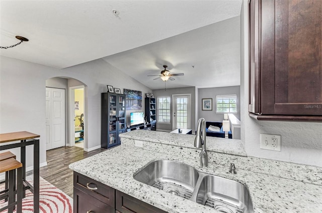 kitchen with arched walkways, dark brown cabinetry, a sink, and light stone counters