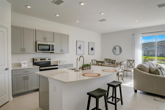 kitchen with sink, an island with sink, a kitchen bar, gray cabinetry, and stainless steel appliances