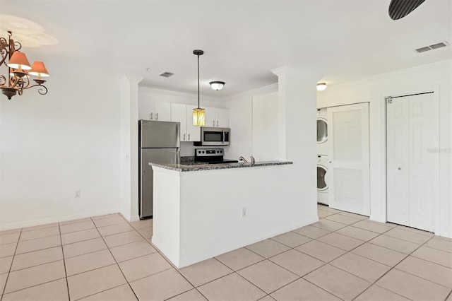 kitchen with appliances with stainless steel finishes, white cabinetry, dark stone counters, stacked washer / drying machine, and pendant lighting