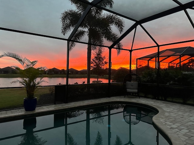 pool at dusk featuring glass enclosure, a patio area, and a water view