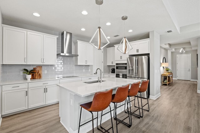 kitchen with wall chimney range hood, a kitchen island with sink, pendant lighting, white cabinets, and appliances with stainless steel finishes