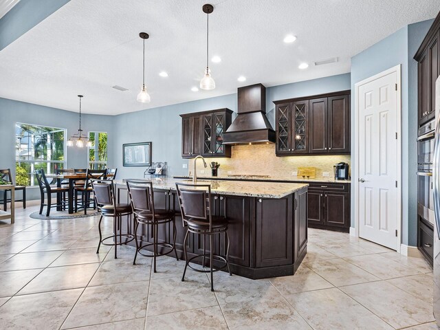 kitchen with hanging light fixtures, custom exhaust hood, a kitchen island with sink, backsplash, and dark brown cabinets