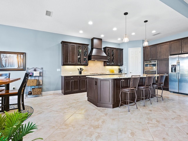 kitchen with appliances with stainless steel finishes, custom range hood, dark brown cabinetry, pendant lighting, and an island with sink