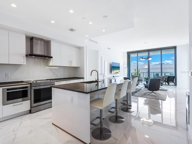 kitchen with wall chimney range hood, a center island with sink, backsplash, white cabinetry, and stainless steel appliances