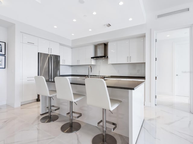 kitchen featuring wall chimney exhaust hood, stainless steel refrigerator, a kitchen bar, and white cabinets