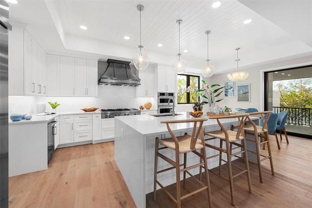 kitchen with a raised ceiling, an island with sink, white cabinets, custom exhaust hood, and light wood-type flooring