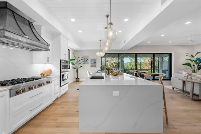 kitchen featuring sink, premium range hood, a large island with sink, decorative light fixtures, and white cabinets