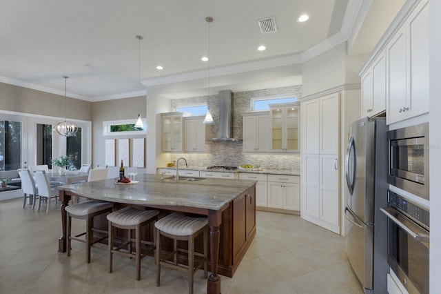 kitchen featuring light stone countertops, wall chimney range hood, an island with sink, pendant lighting, and appliances with stainless steel finishes