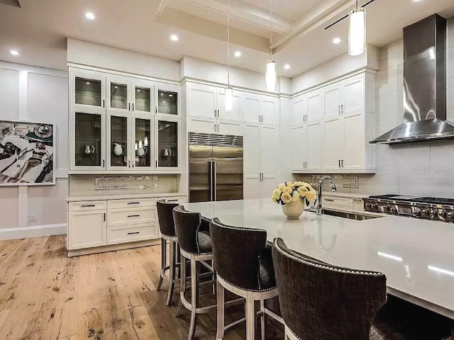 kitchen with wall chimney range hood, a kitchen breakfast bar, light hardwood / wood-style floors, decorative light fixtures, and built in refrigerator