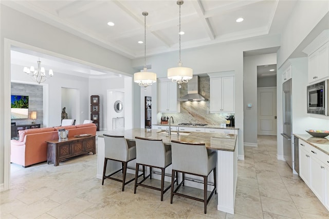 kitchen with light stone counters, white cabinets, an island with sink, pendant lighting, and wall chimney exhaust hood