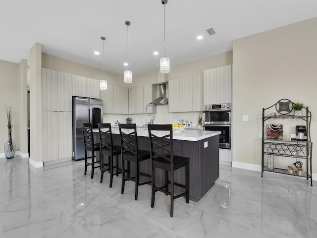 kitchen with decorative light fixtures, a center island with sink, wall chimney exhaust hood, appliances with stainless steel finishes, and a breakfast bar