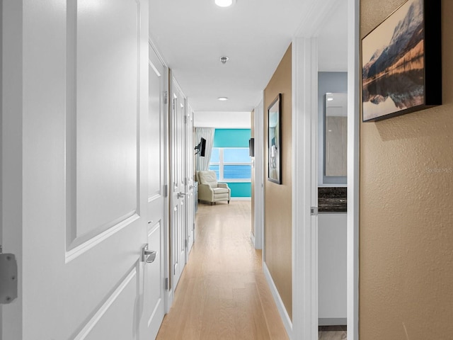 hall featuring a textured wall, light wood-type flooring, and baseboards