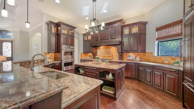 kitchen with arched walkways, light stone countertops, a sink, double oven, and an island with sink
