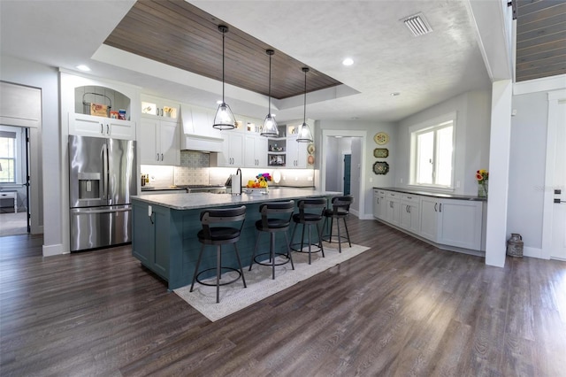 kitchen with a center island with sink, a tray ceiling, stainless steel fridge, pendant lighting, and white cabinets