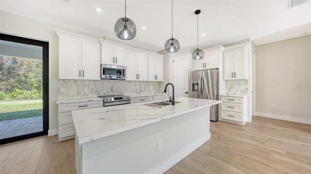 kitchen with an island with sink, stainless steel appliances, pendant lighting, light wood-type flooring, and white cabinetry