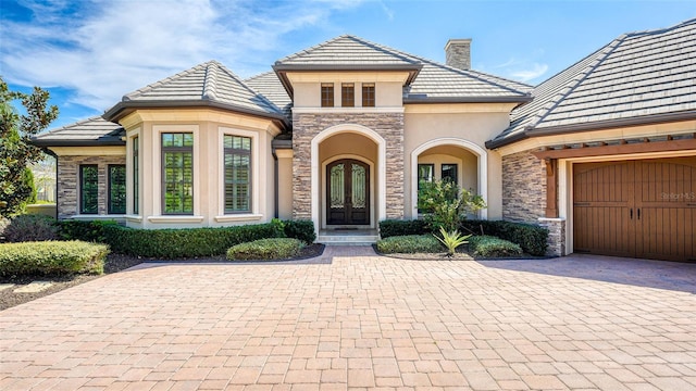 french country home featuring stucco siding, french doors, a chimney, decorative driveway, and stone siding