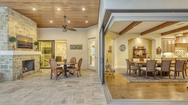 dining space with beamed ceiling, a stone fireplace, ceiling fan, and wooden ceiling