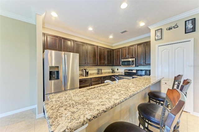 kitchen with a breakfast bar area, stainless steel appliances, ornamental molding, light stone countertops, and dark brown cabinetry