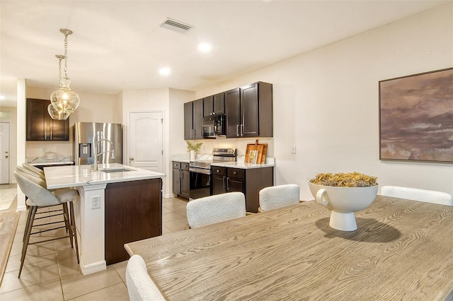 kitchen featuring hanging light fixtures, stainless steel appliances, a center island with sink, dark brown cabinetry, and light tile patterned floors