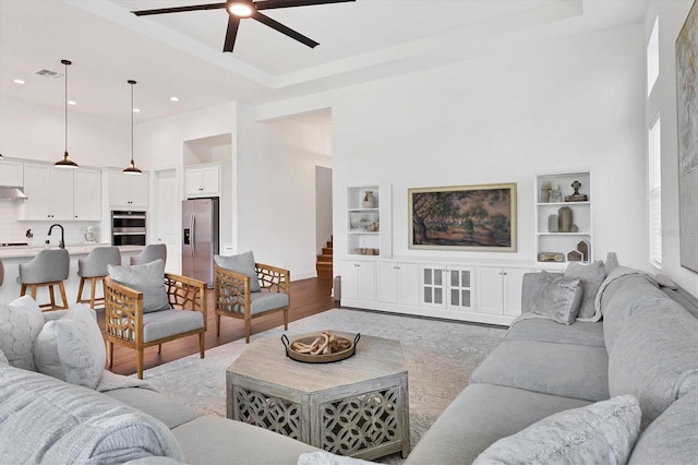 living room with ceiling fan, a tray ceiling, a high ceiling, built in shelves, and light hardwood / wood-style floors