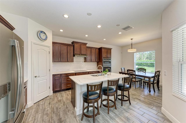 kitchen featuring hanging light fixtures, an island with sink, appliances with stainless steel finishes, light stone countertops, and light hardwood / wood-style floors