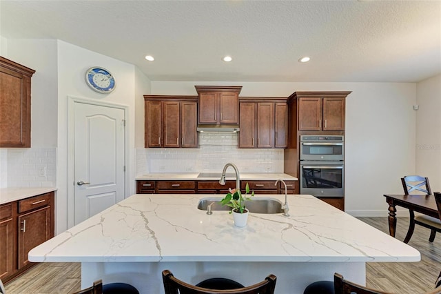 kitchen with stainless steel double oven, a center island with sink, sink, a kitchen bar, and light wood-type flooring