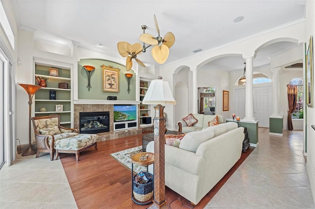 living room featuring a tile fireplace, ceiling fan, built in shelves, ornamental molding, and light tile patterned floors