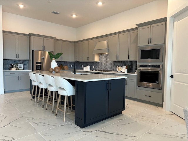 kitchen featuring appliances with stainless steel finishes, custom exhaust hood, decorative backsplash, and a kitchen island with sink