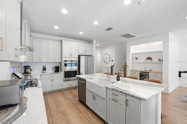 kitchen featuring stainless steel appliances, light stone countertops, beverage cooler, a kitchen island with sink, and light hardwood / wood-style flooring