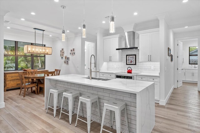 kitchen with hanging light fixtures, wall chimney range hood, a kitchen island with sink, white cabinets, and sink