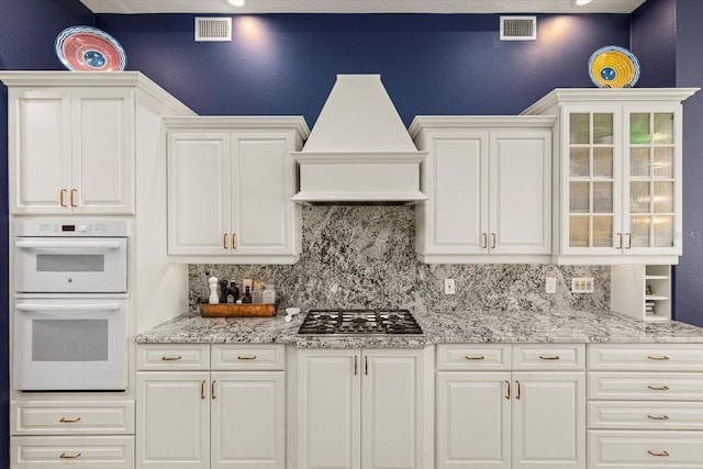 kitchen with light stone counters, white cabinets, stainless steel gas cooktop, custom range hood, and white double oven