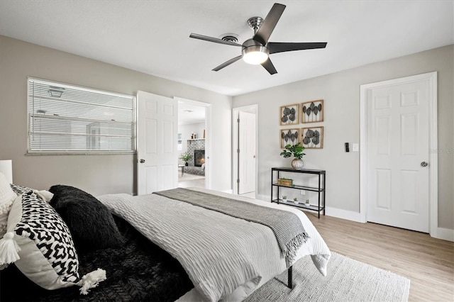 bedroom featuring baseboards, a stone fireplace, light wood-type flooring, and a ceiling fan