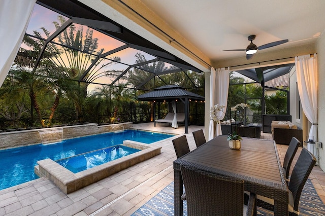 pool at dusk featuring a lanai, ceiling fan, a gazebo, pool water feature, and an in ground hot tub