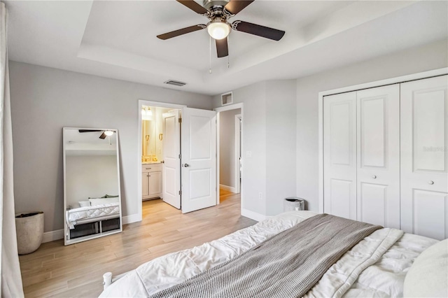 bedroom with ensuite bathroom, light wood-type flooring, a raised ceiling, a closet, and ceiling fan