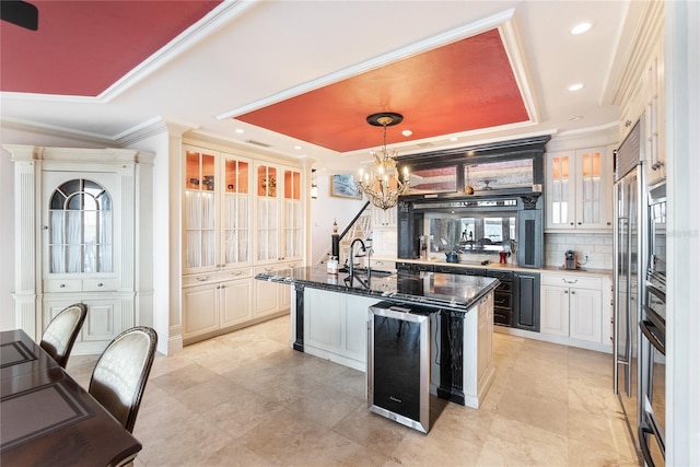 kitchen with an island with sink, backsplash, a tray ceiling, pendant lighting, and beverage cooler