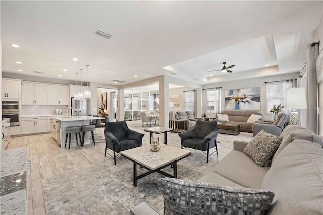 living room featuring sink, light hardwood / wood-style flooring, a tray ceiling, and ceiling fan with notable chandelier