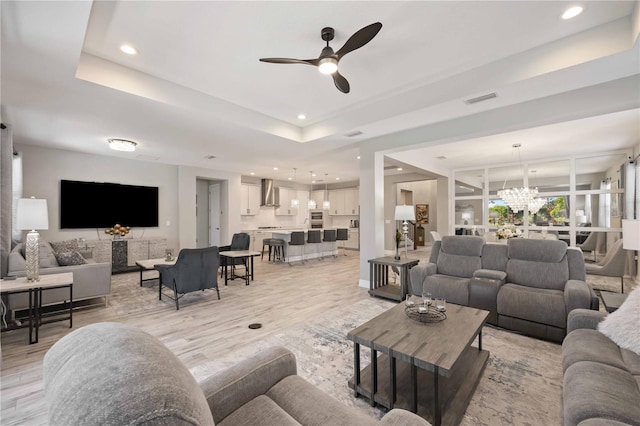 living room with ceiling fan with notable chandelier, light wood-type flooring, and a raised ceiling