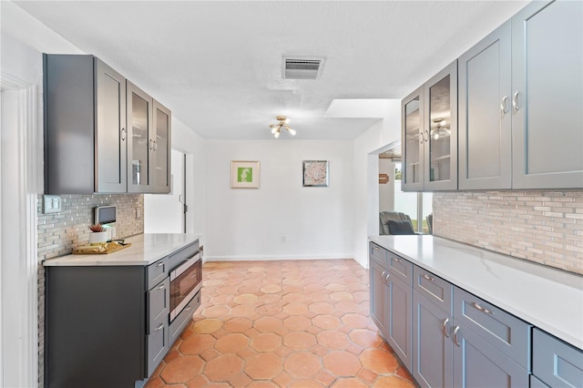 kitchen featuring glass insert cabinets, light countertops, gray cabinetry, visible vents, and decorative backsplash