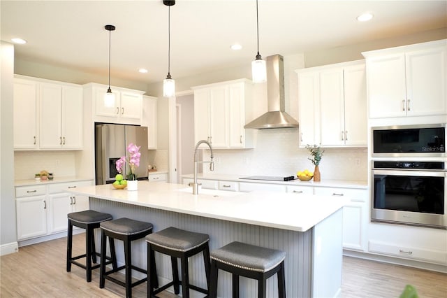 kitchen with white cabinets, a center island with sink, appliances with stainless steel finishes, light wood-type flooring, and wall chimney exhaust hood