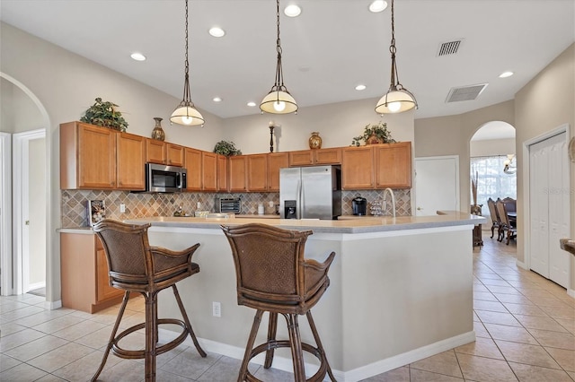 kitchen featuring tasteful backsplash, hanging light fixtures, stainless steel appliances, light tile patterned floors, and a kitchen island with sink