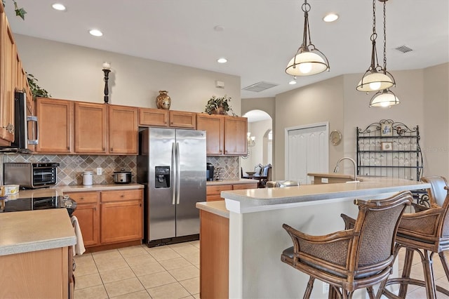 kitchen with light tile patterned flooring, an island with sink, hanging light fixtures, stainless steel appliances, and decorative backsplash