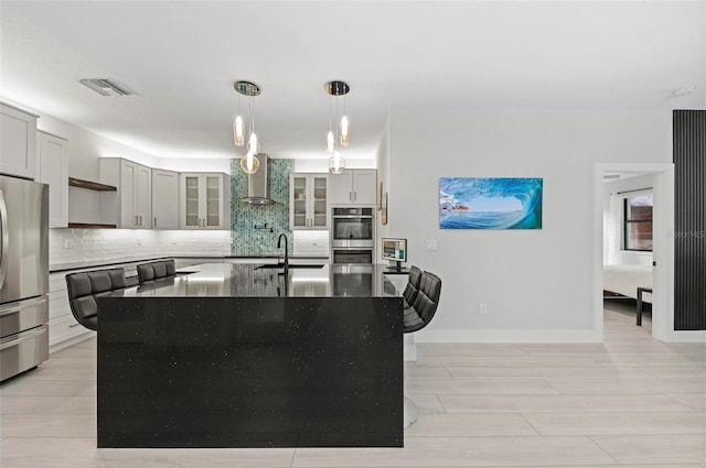 kitchen with dark stone counters, wall chimney exhaust hood, stainless steel appliances, a kitchen island with sink, and sink