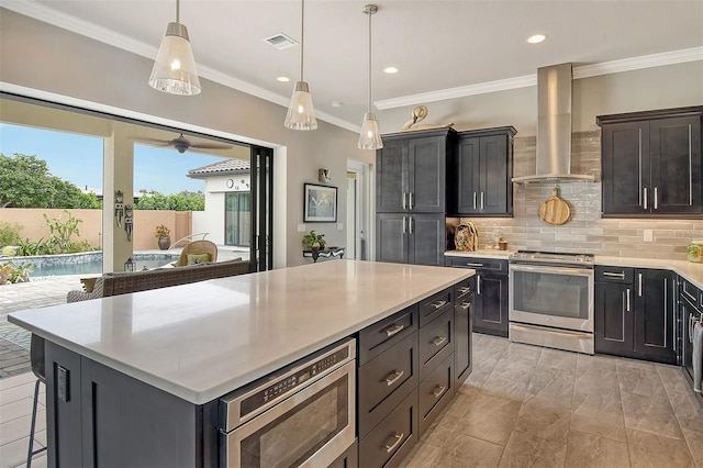 kitchen featuring appliances with stainless steel finishes, a center island, hanging light fixtures, and wall chimney range hood