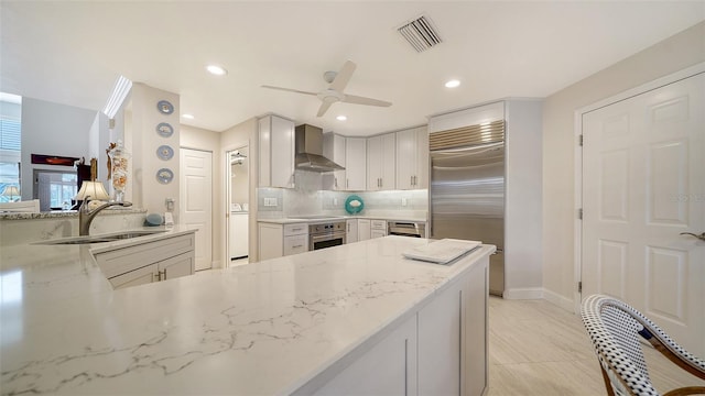 kitchen featuring wall chimney range hood, kitchen peninsula, light stone counters, sink, and stainless steel appliances