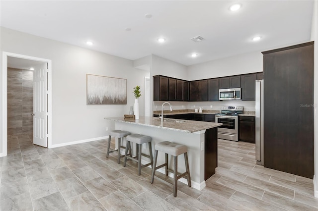 kitchen featuring dark brown cabinets, an island with sink, appliances with stainless steel finishes, a kitchen bar, and sink